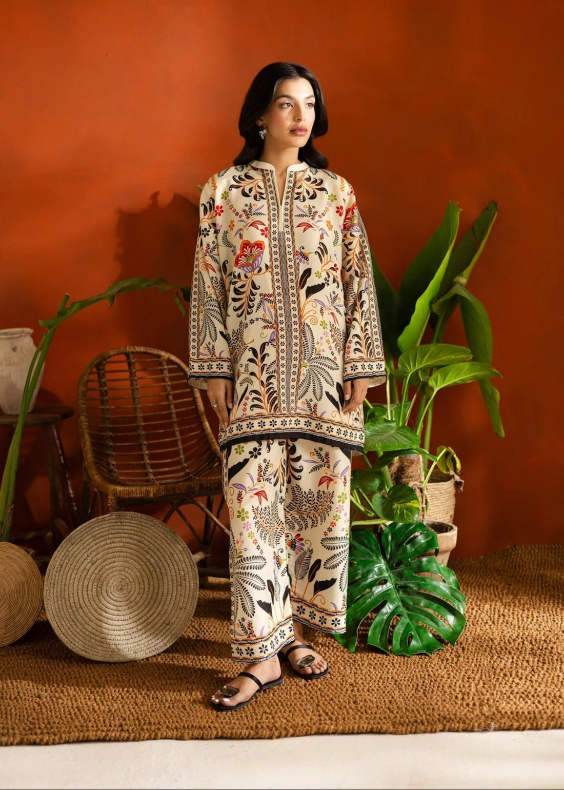 Woman in a patterned outfit standing in a room with plants and a red wall.