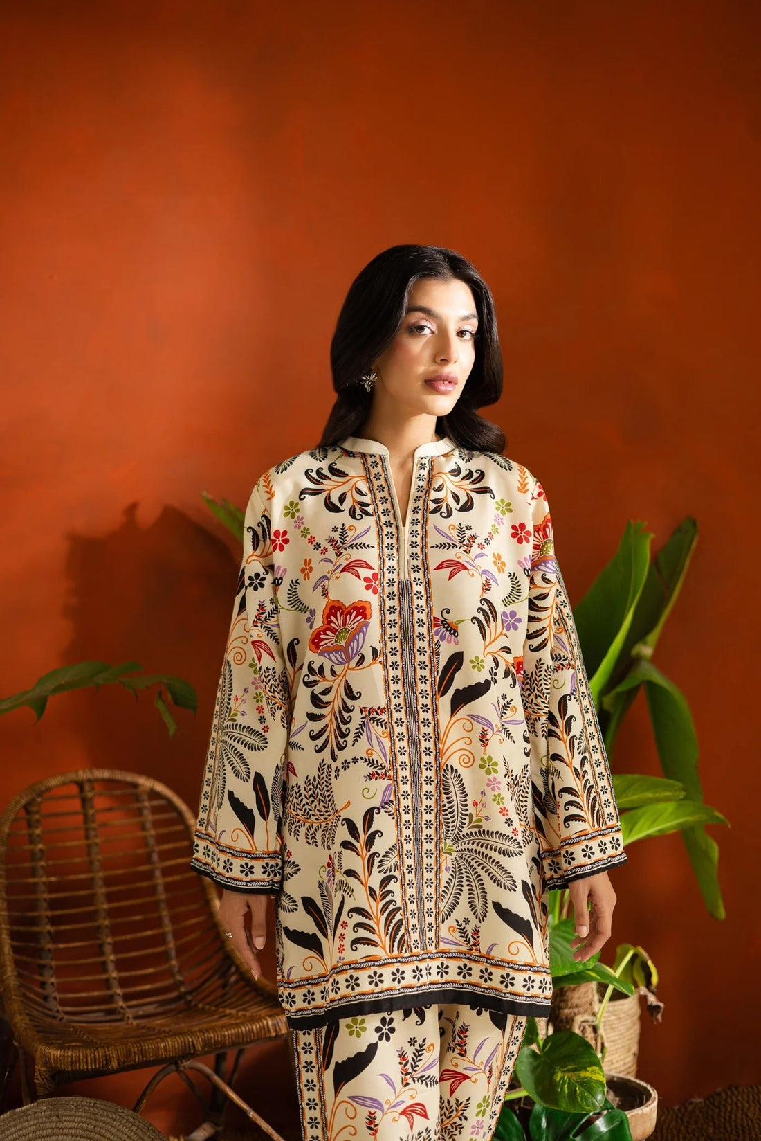 Woman in a patterned outfit standing in a room with plants and a red wall.